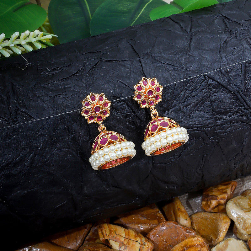 A pair of ornate jhumka earrings with red and gold detailing, adorned with small white pearls, displayed on a textured black surface with green leaves in the background.