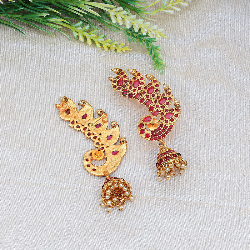 Two ornate gold earrings with red gemstone accents and dangling pearl-like beads, displayed on a light fabric background with green foliage.