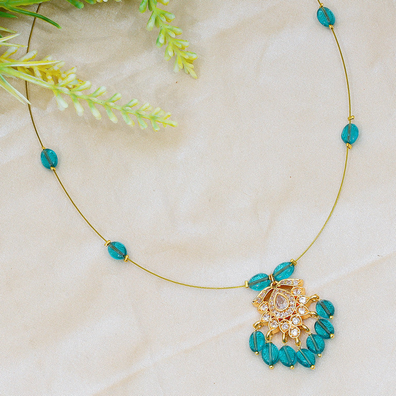 A delicate necklace with a gold chain featuring turquoise beads and an ornate gold pendant with clear gemstones, displayed on a light fabric background with green foliage.