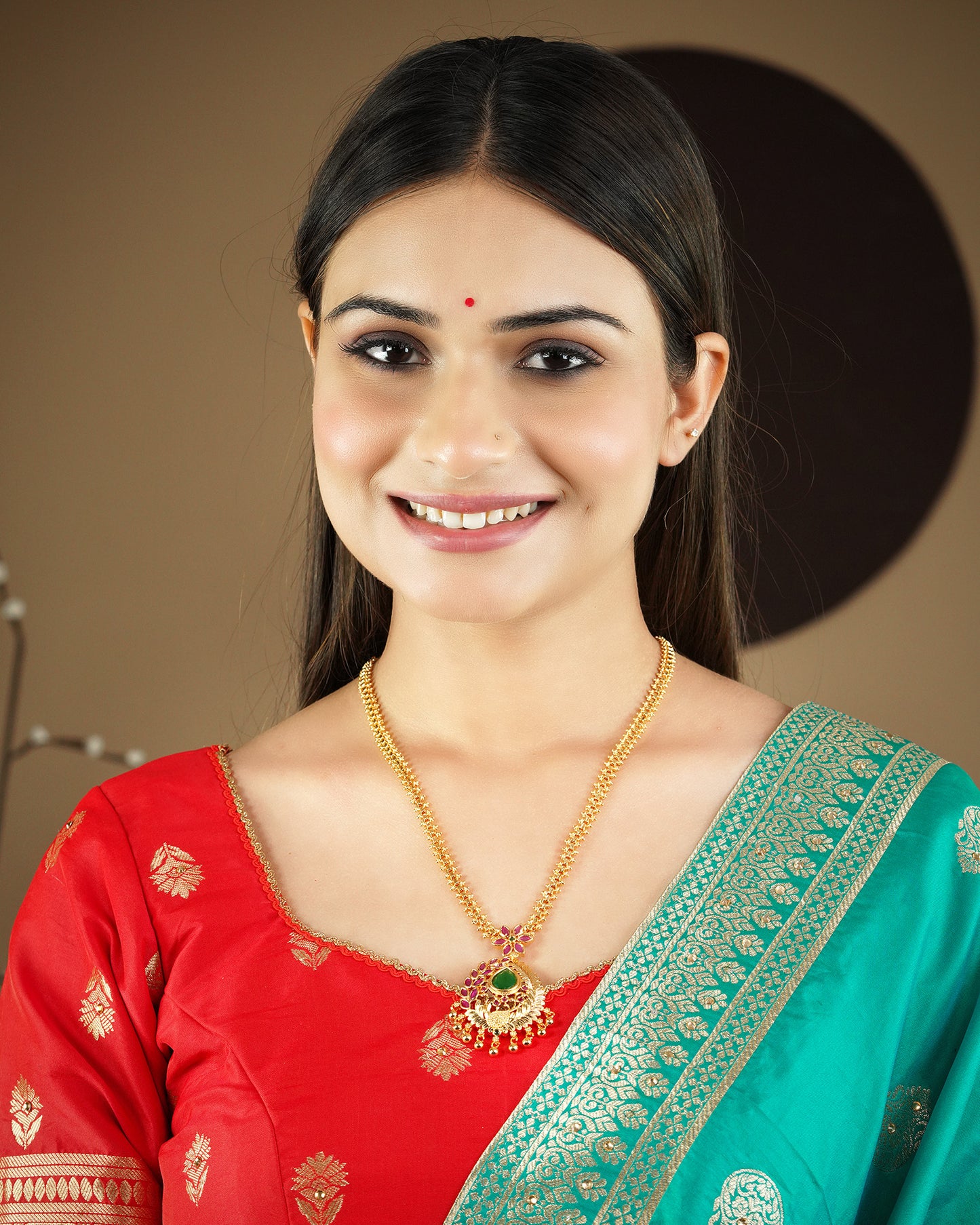 A woman wearing a red and green traditional outfit and a gold necklace with a green pendant, smiling.