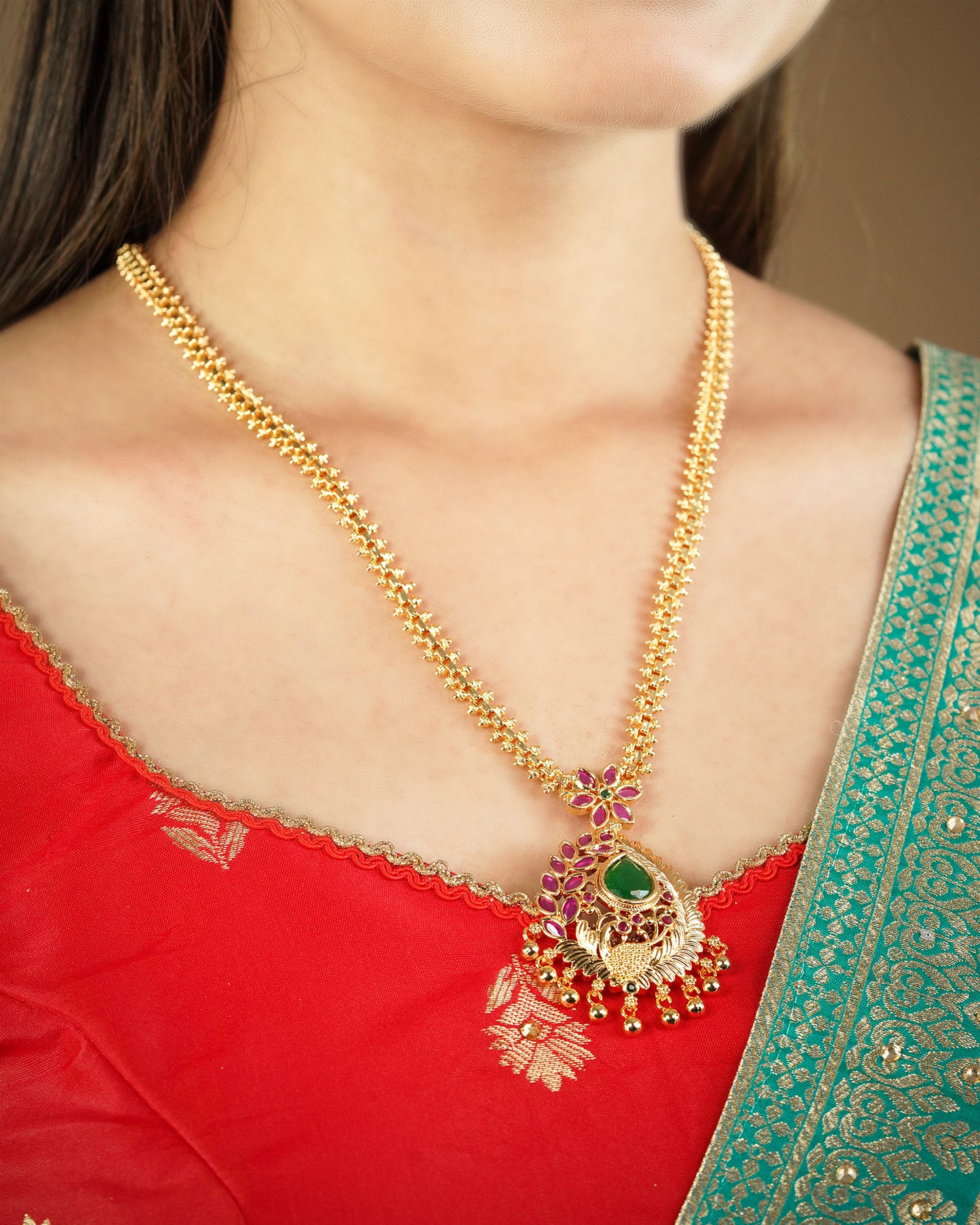 A person wearing a gold necklace with a green and purple pendant, dressed in a red and green traditional outfit.