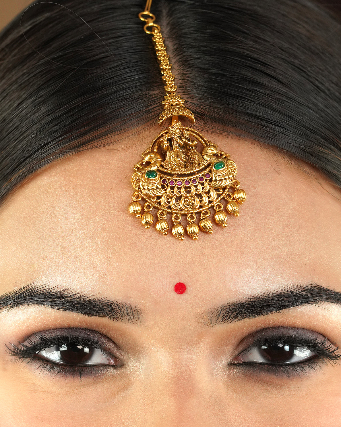 A close-up of a person wearing a gold maang tikka with intricate designs and colorful gemstones.