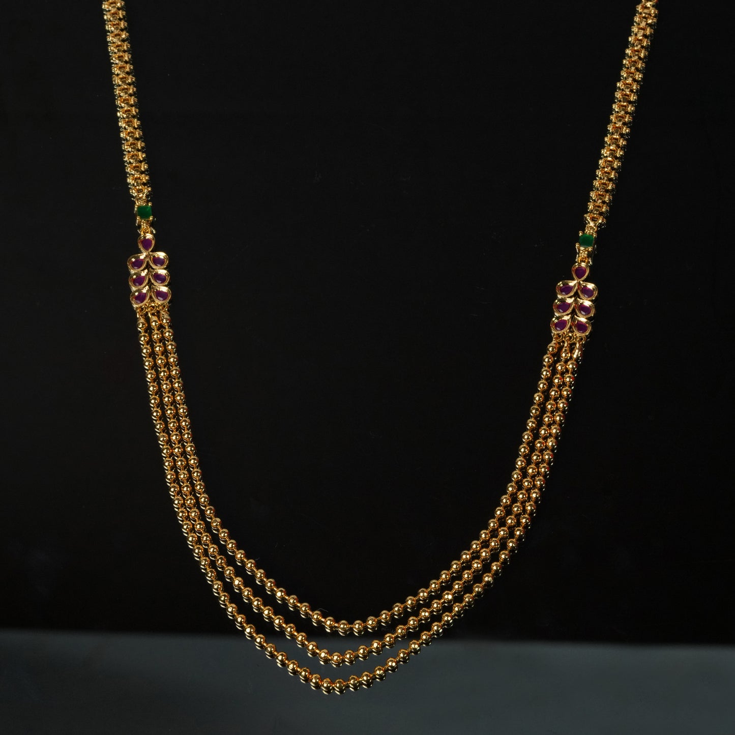 A gold necklace with three strands, featuring intricate designs and red and green gemstone accents.