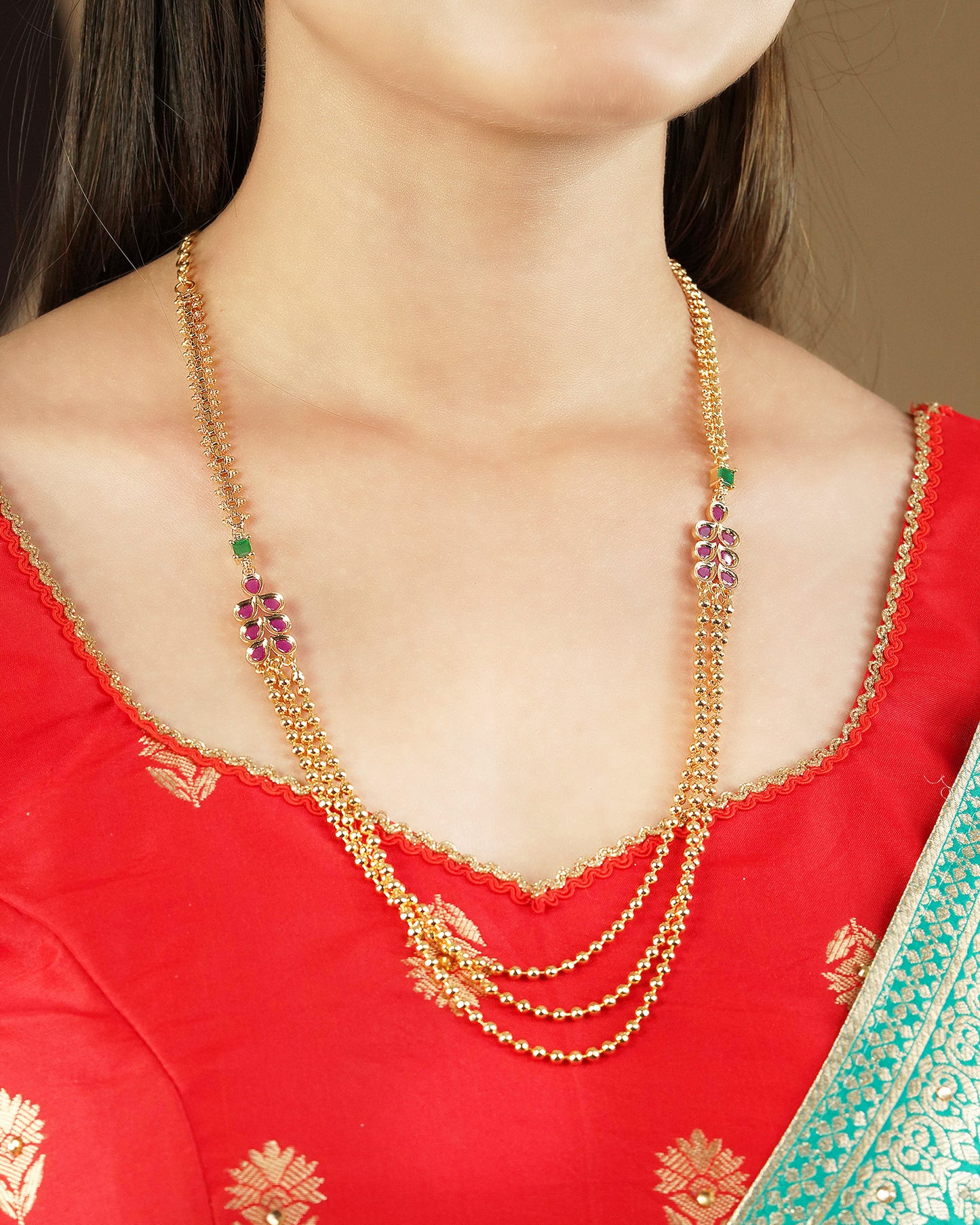 A woman wearing a gold layered necklace with green and pink gemstone accents, paired with a red traditional outfit.