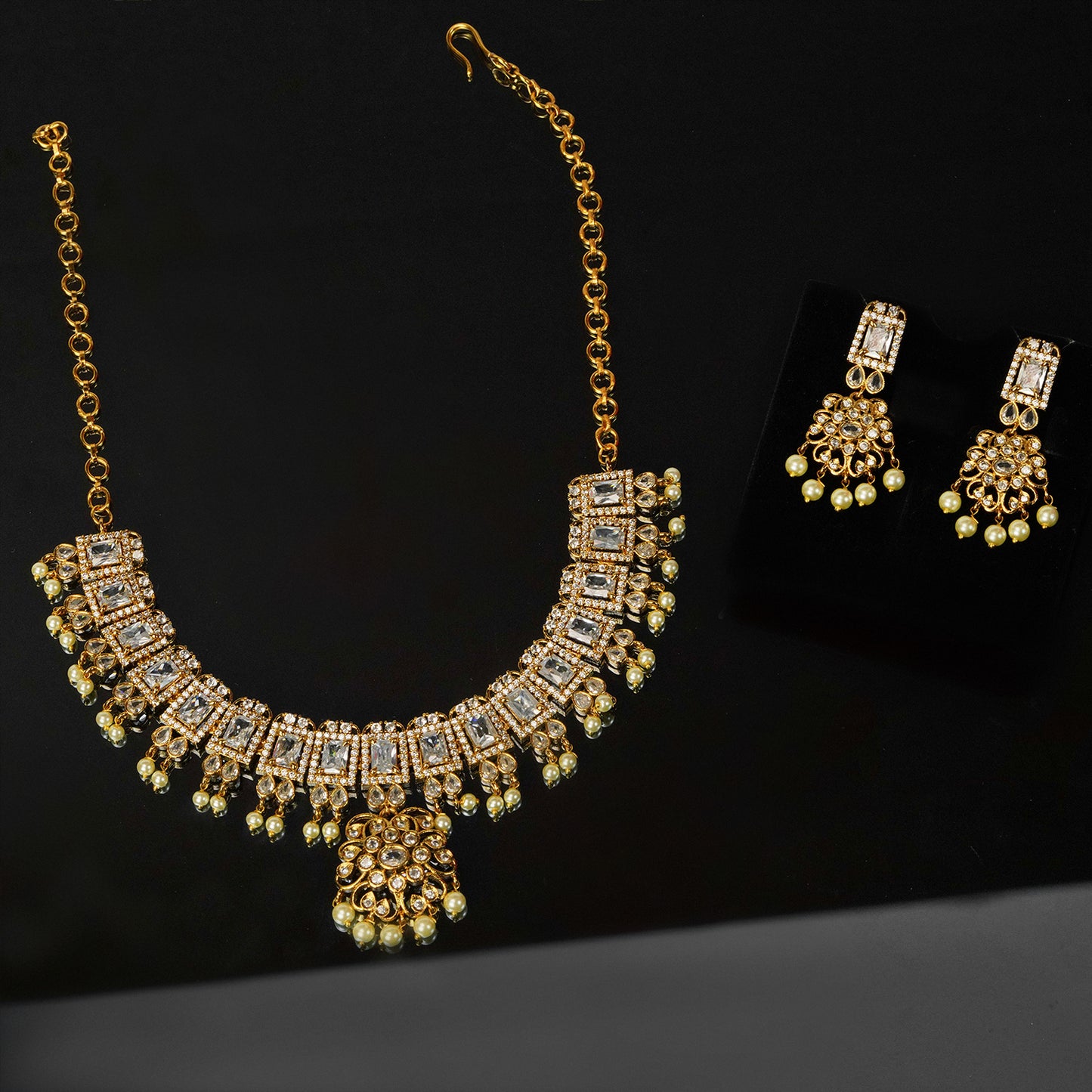 A gold necklace with intricate square and circular designs, adorned with white and green gemstones, accompanied by matching earrings.