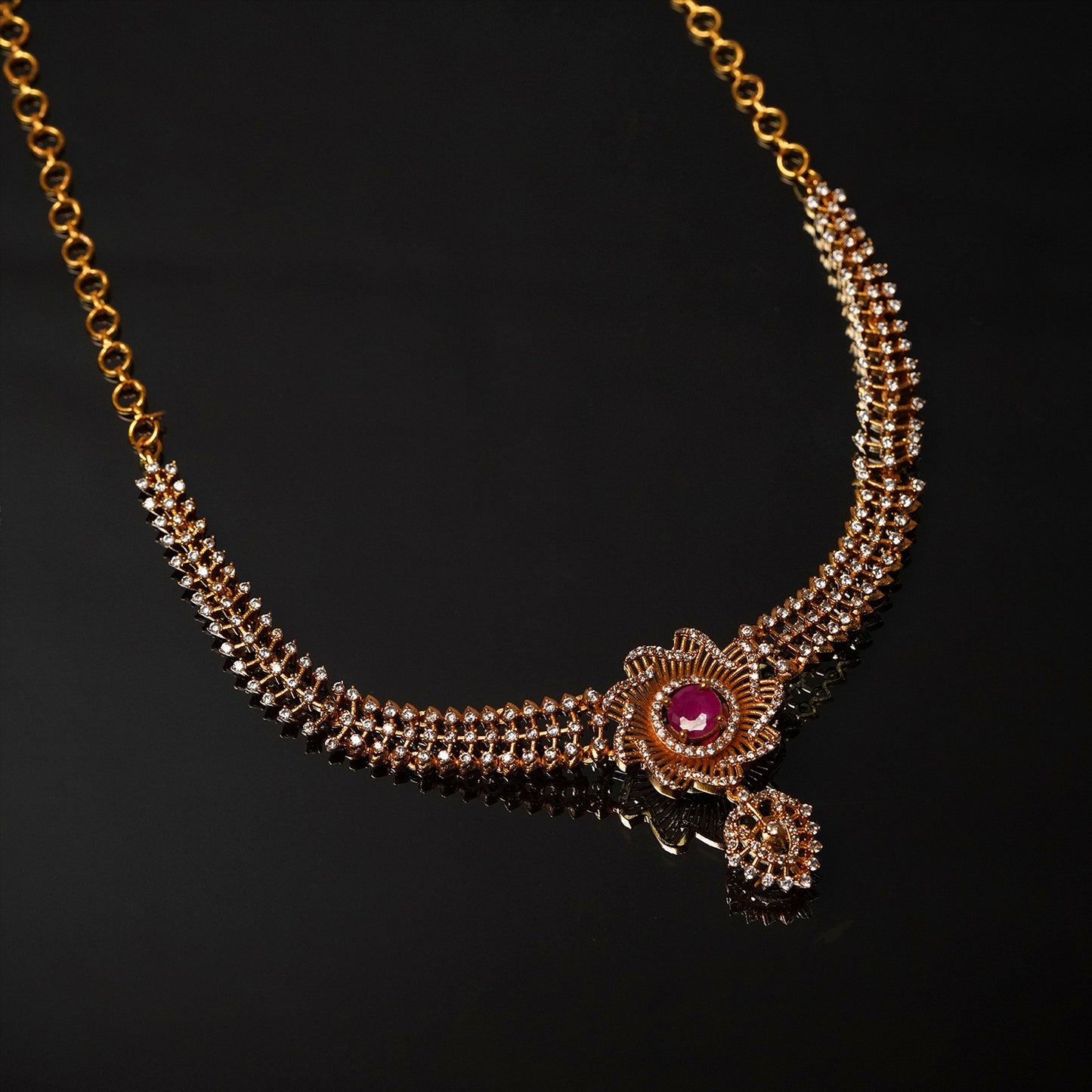 A gold necklace with intricate detailing, featuring a central red gemstone and multiple small white stones.