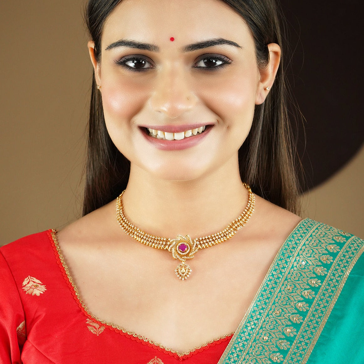 A woman wearing a gold necklace with a central pink gemstone and matching earrings, dressed in a red and green traditional outfit.