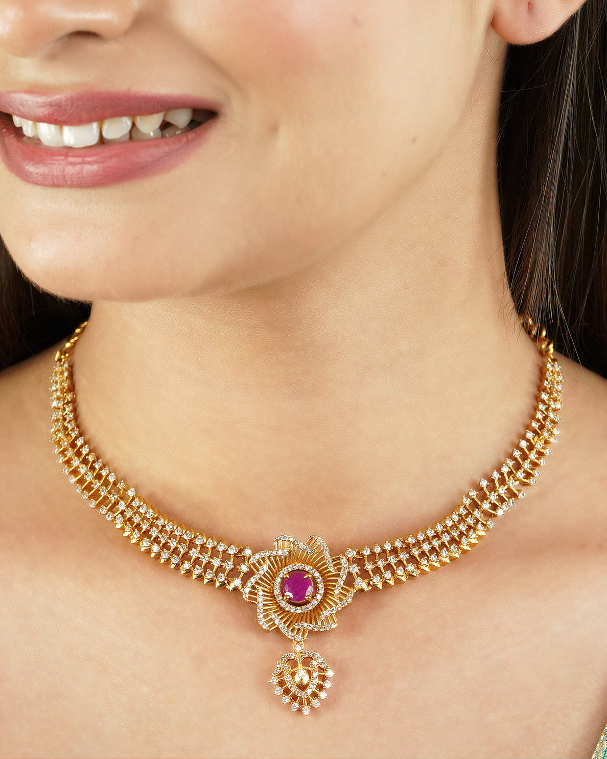 A woman wearing a gold necklace with a central floral design featuring a pink gemstone.