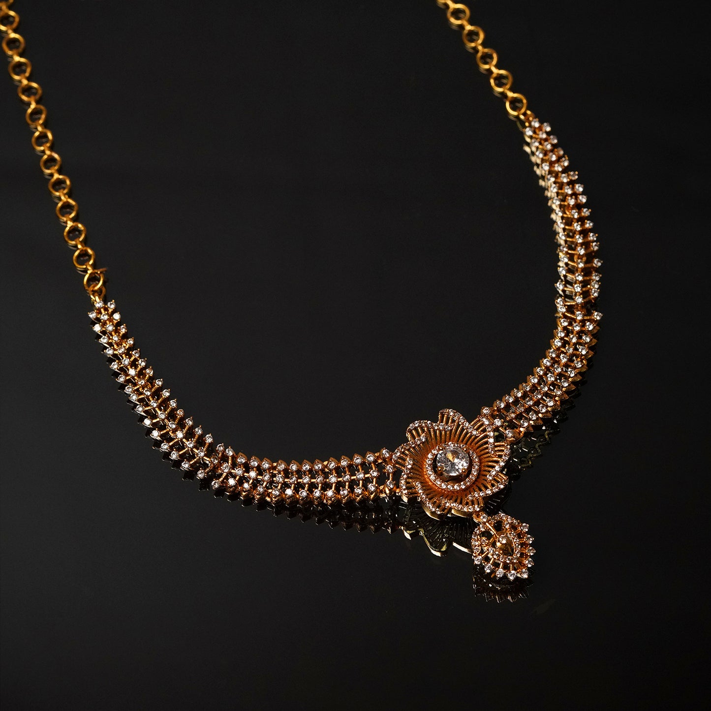 A gold necklace with intricate detailing, featuring a central floral design adorned with a gemstone and a small dangling pendant.