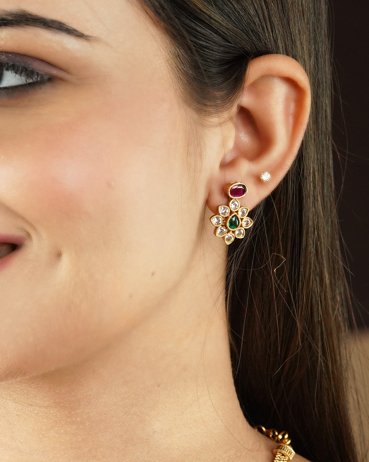 Close-up of a woman's ear wearing a gold floral earring with green and white stones, along with a small stud earring.
