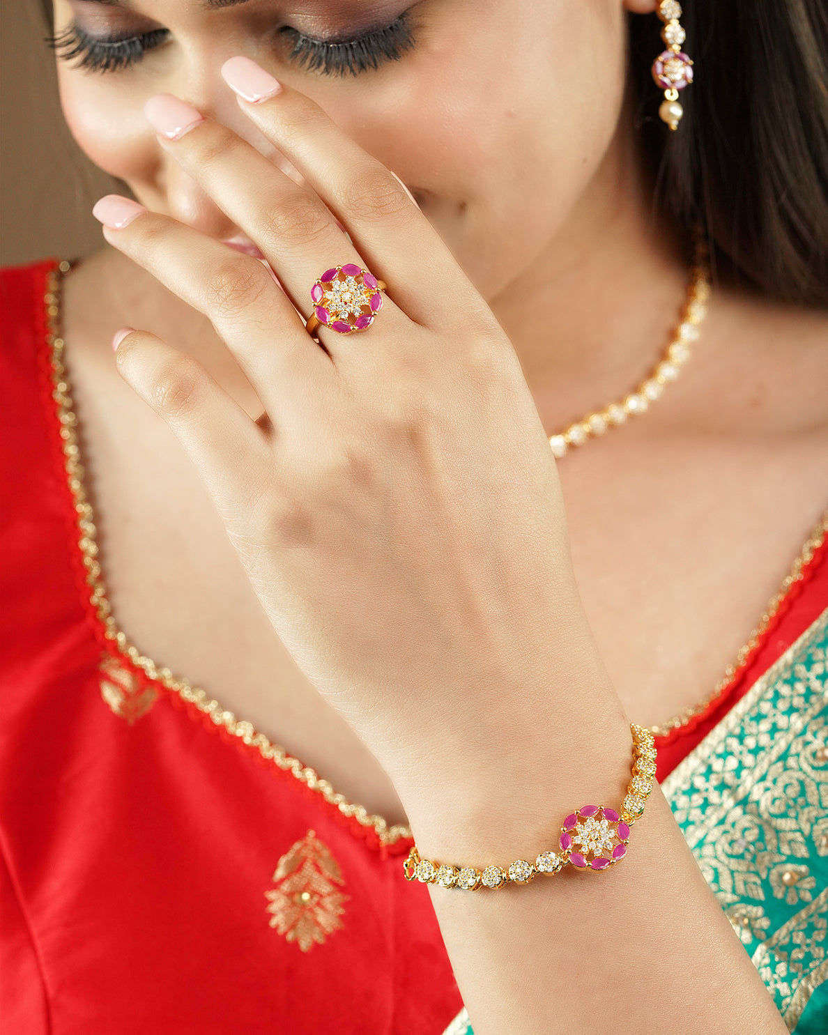 A woman wearing a red and green traditional outfit, showcasing a gold bracelet and ring set adorned with pink and white stones.