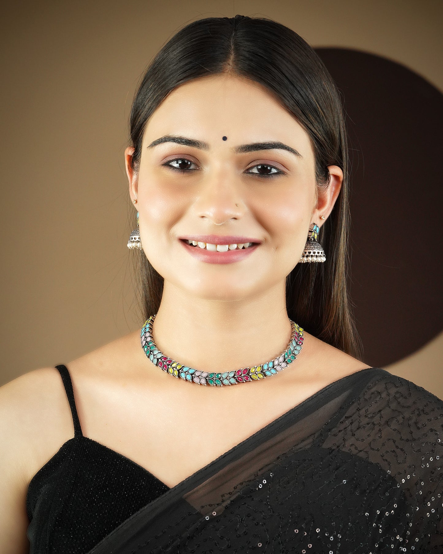 A woman wearing a colorful beaded choker necklace and silver jhumka earrings, dressed in a black saree with a sequined border.
