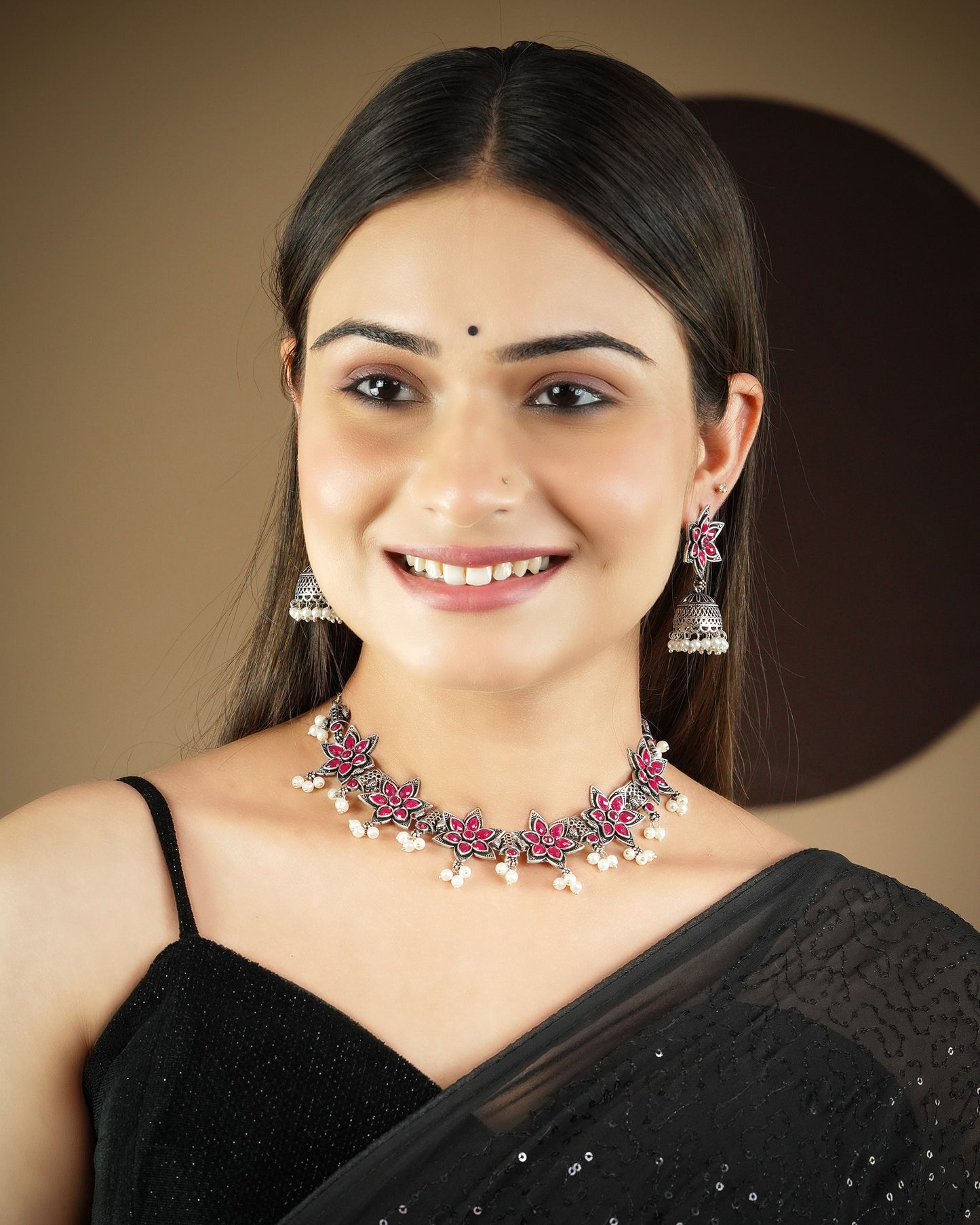 A woman wearing a black saree and a matching set of jewelry, including a floral necklace with pink and white accents and dangling earrings.