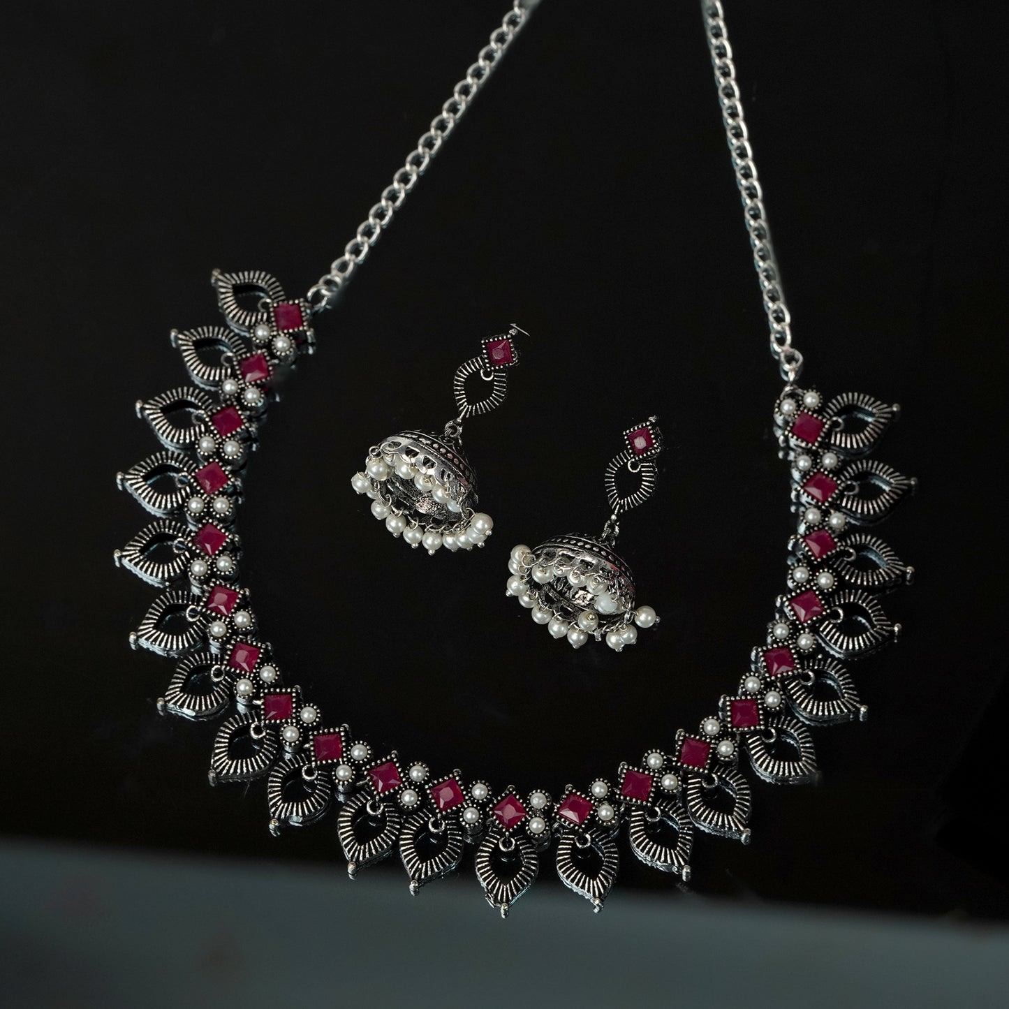 A silver necklace with intricate leaf designs and red gemstone accents, accompanied by matching earrings.