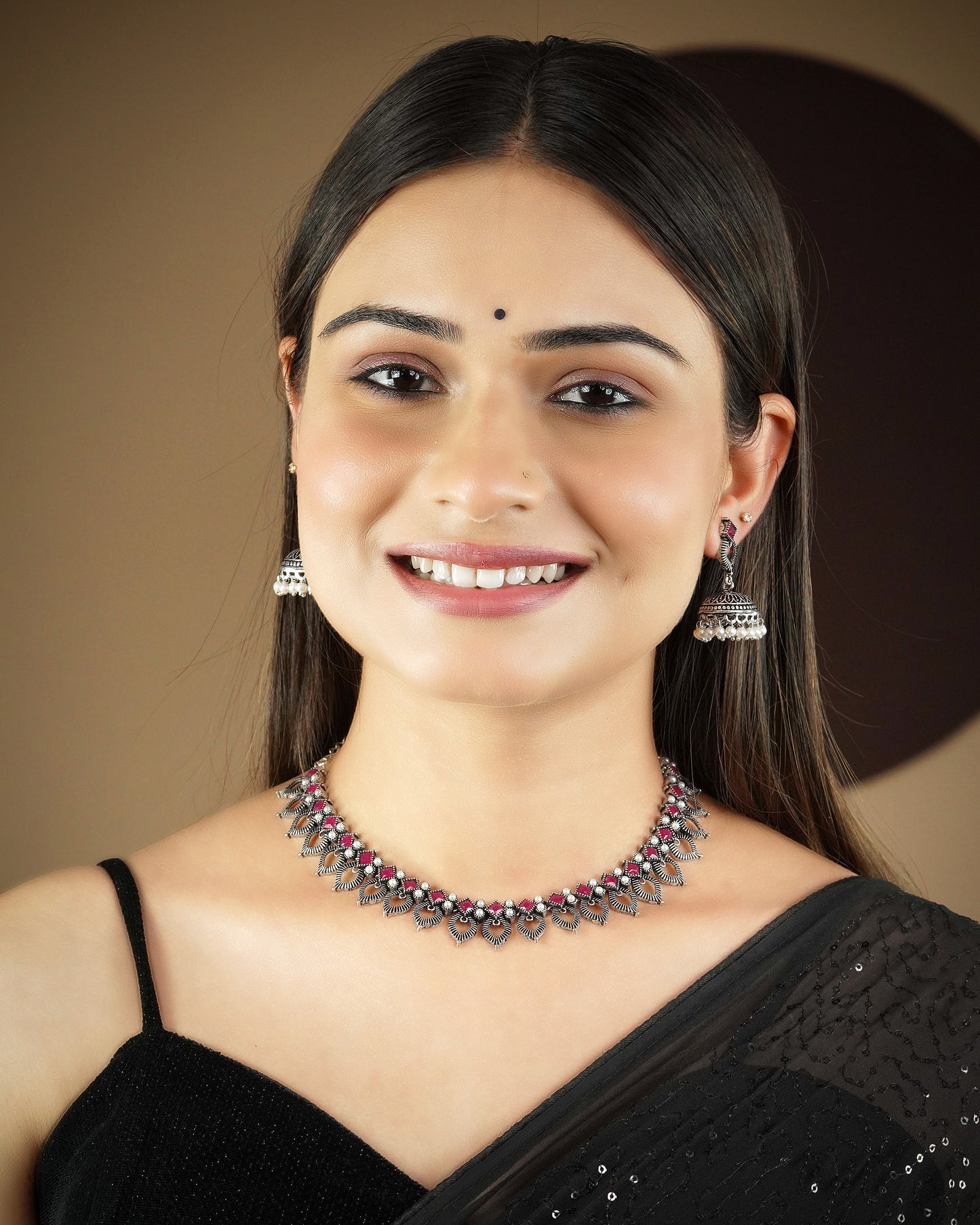 A woman wearing a black saree, silver jhumka earrings, and a silver necklace with red and green stones.
