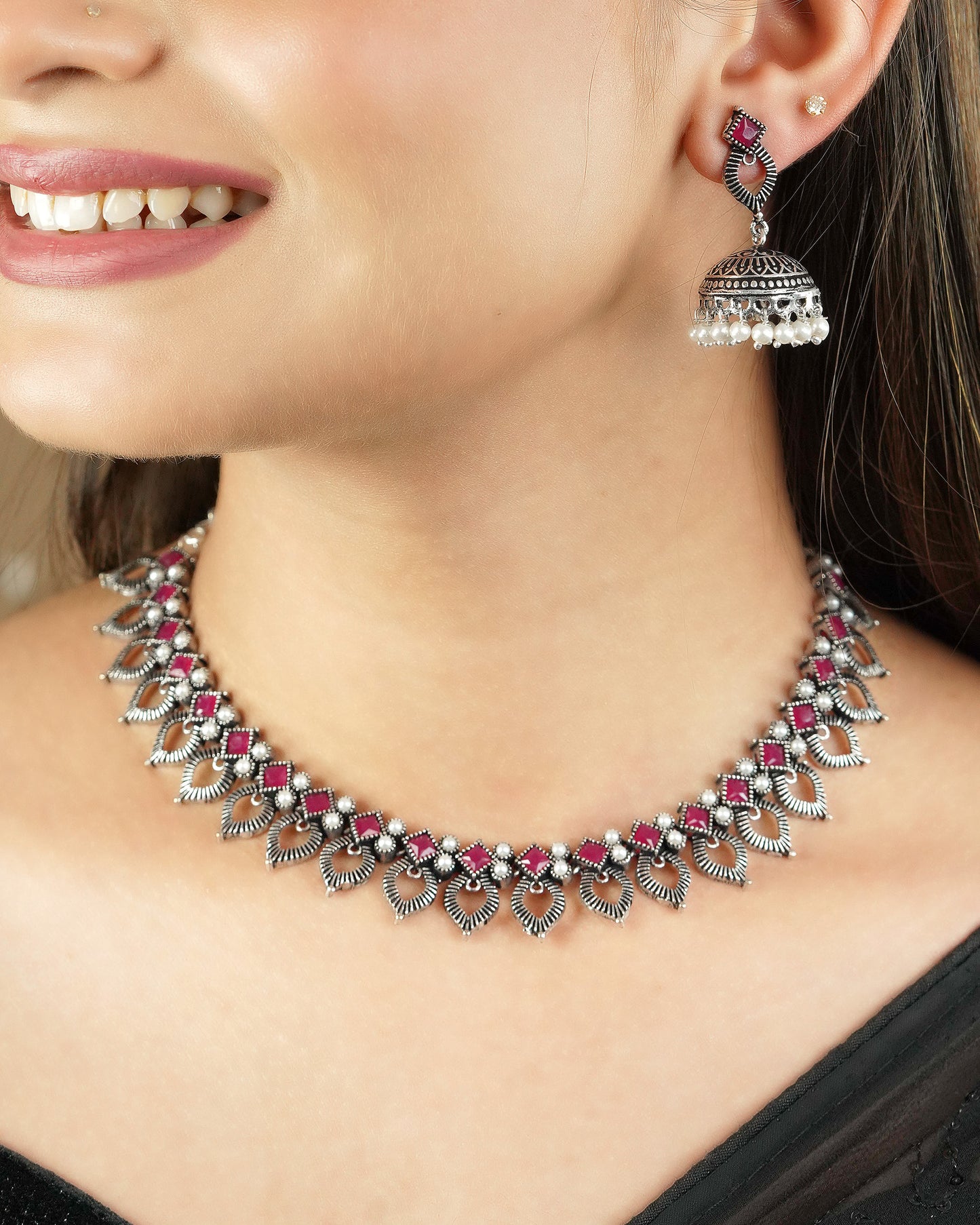 A woman wearing a silver necklace with intricate designs and red gemstone accents, paired with a matching silver earring featuring a jhumka design and pearl drops.