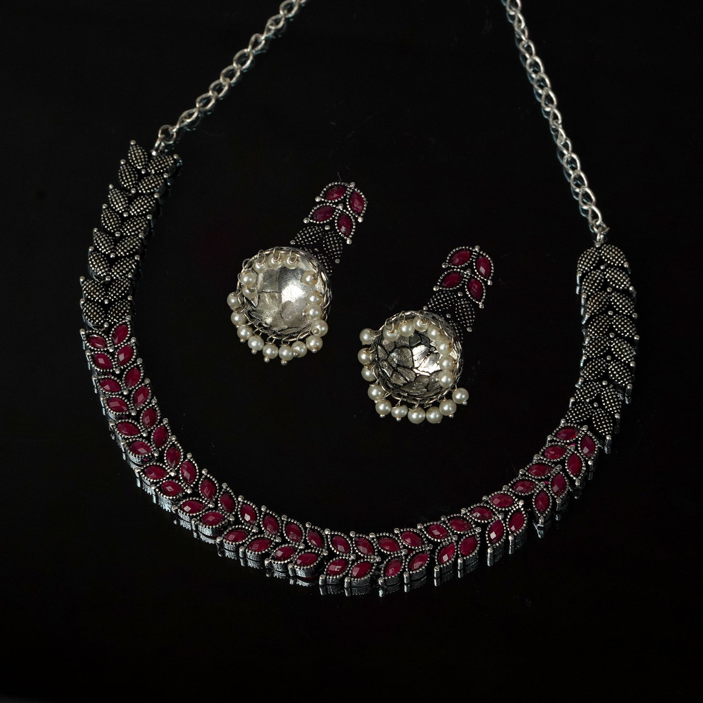 A silver necklace with intricate red and black detailing, accompanied by matching earrings featuring large central stones surrounded by small pearls.