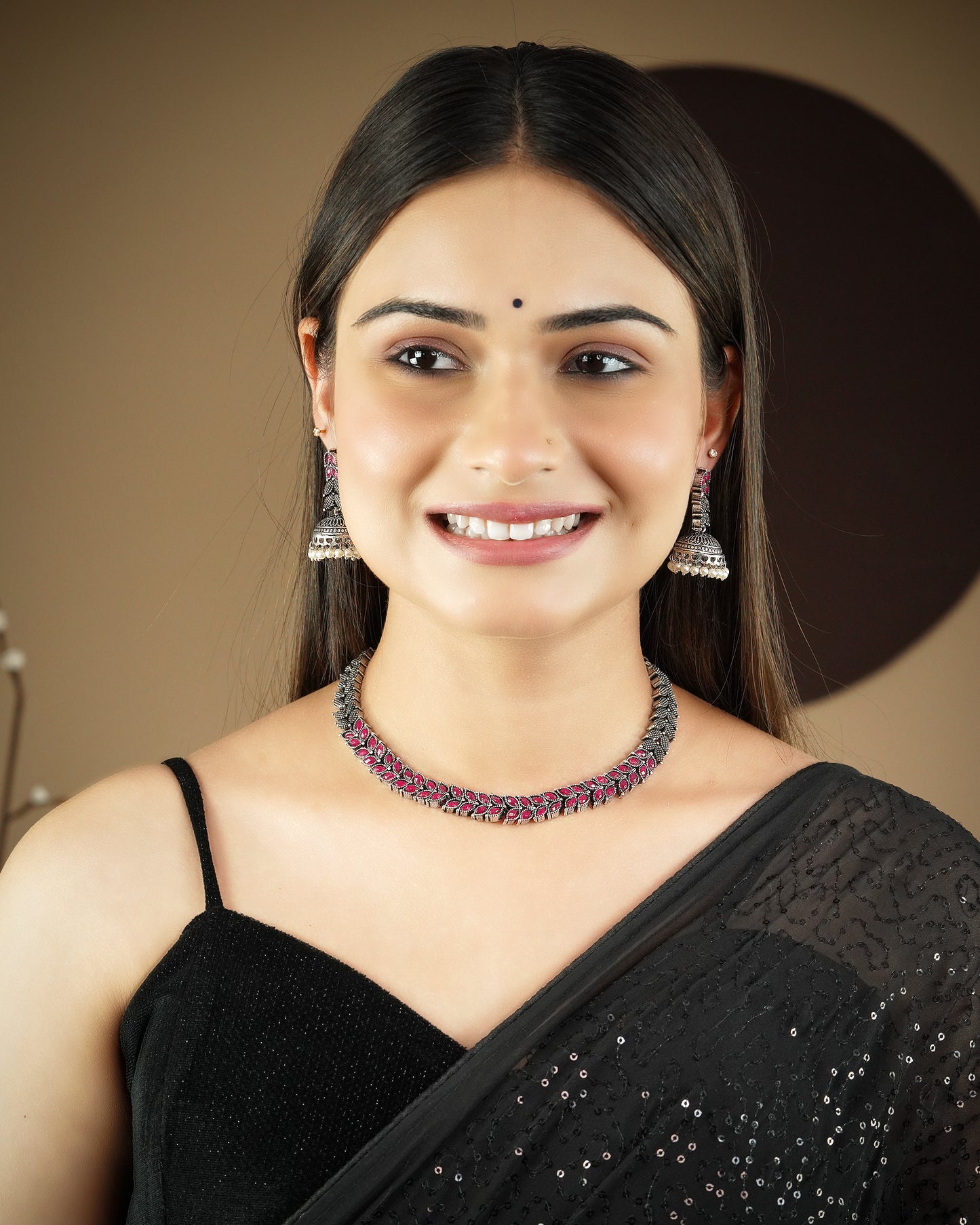 A woman wearing a black saree, silver jhumka earrings, and a silver necklace with pink accents.