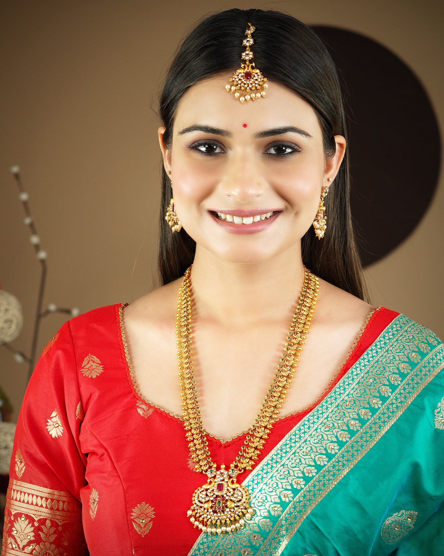 A woman wearing traditional Indian jewelry, including a maang tikka, earrings, and a necklace, paired with a red and green saree.