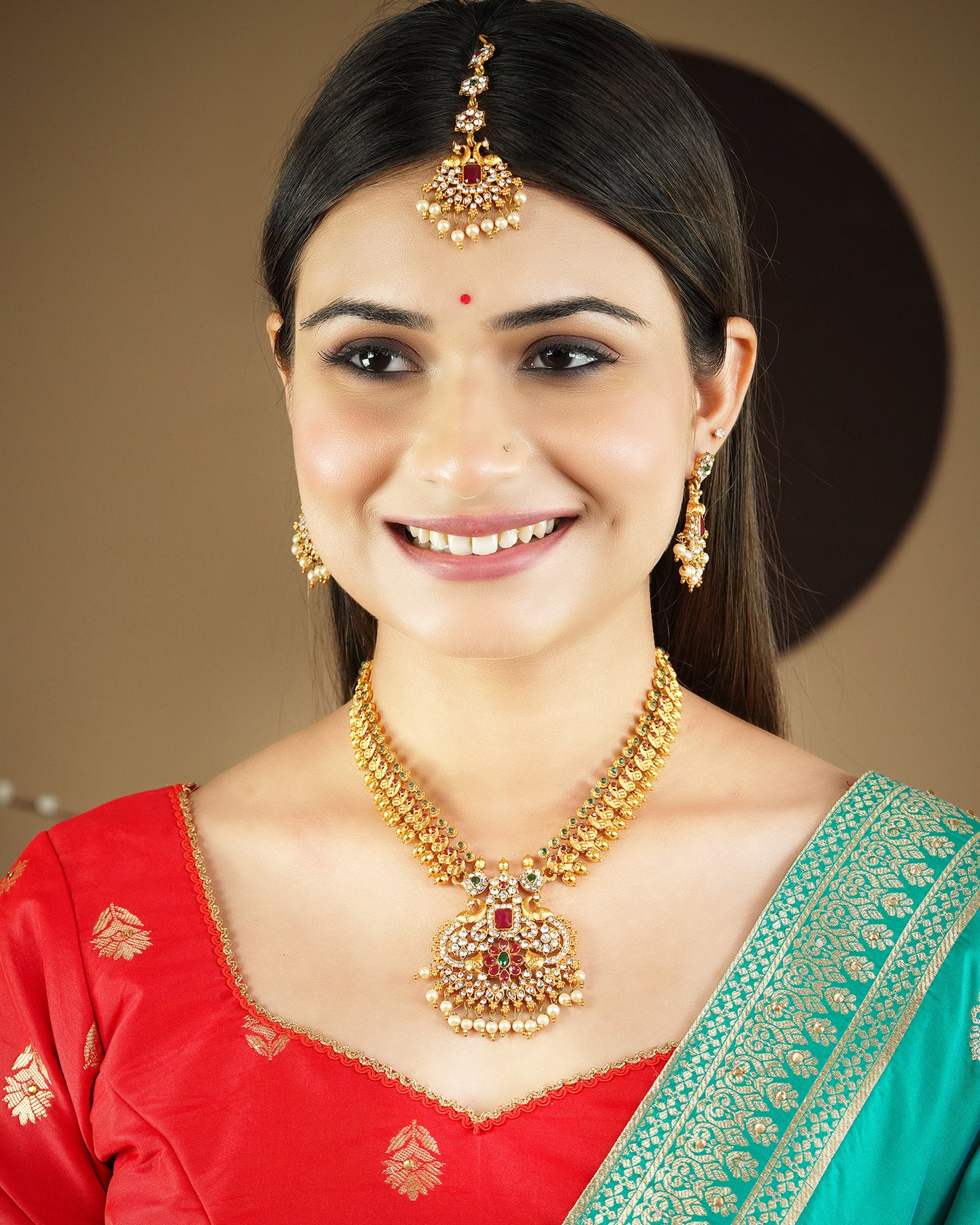 A woman wearing a traditional gold necklace set with matching earrings and a maang tikka, dressed in a red and green saree.