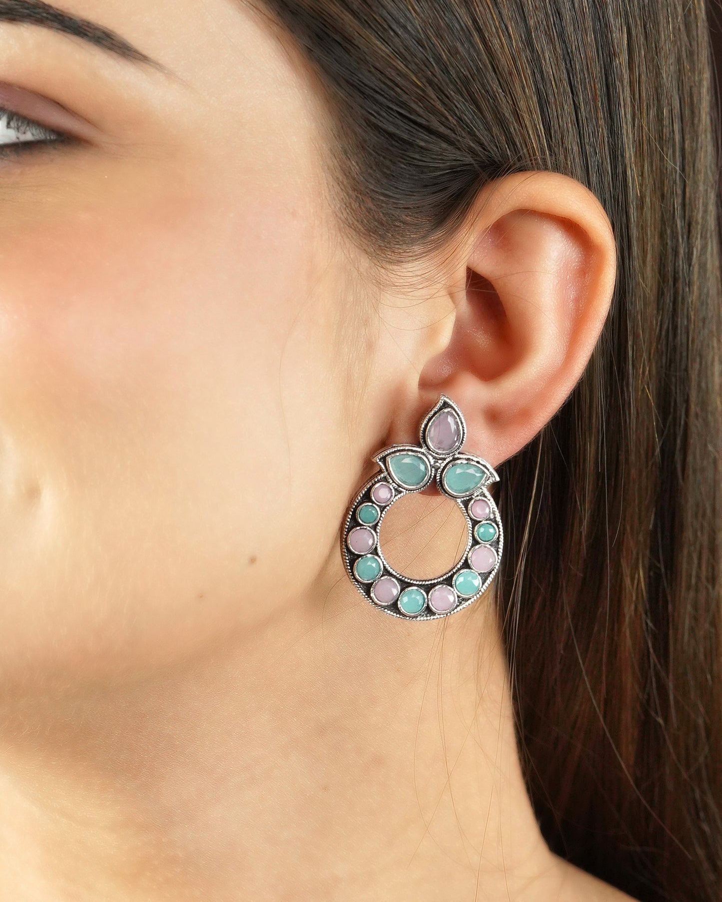 A close-up of a woman's ear wearing a circular earring with pink and green gemstones.