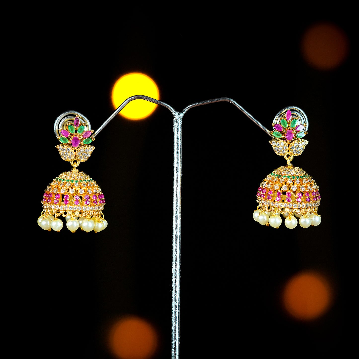 A pair of gold jhumka earrings adorned with pink and green gemstones and pearl drops.