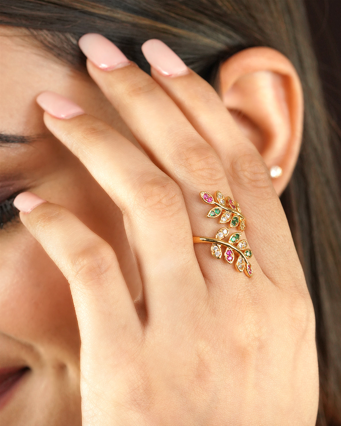 A woman's hand wearing a gold ring with a leaf design adorned with multicolored gemstones.