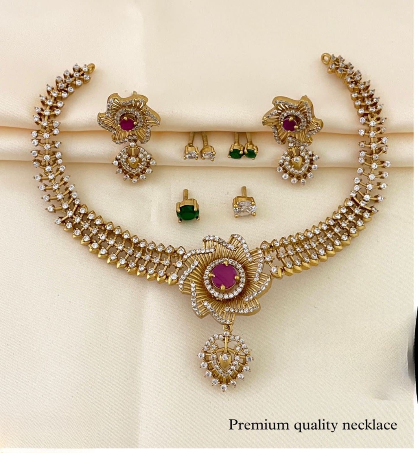 A gold necklace set with intricate floral designs, featuring a central red gemstone and surrounded by smaller white stones, accompanied by matching earrings and additional green and white stone earrings.