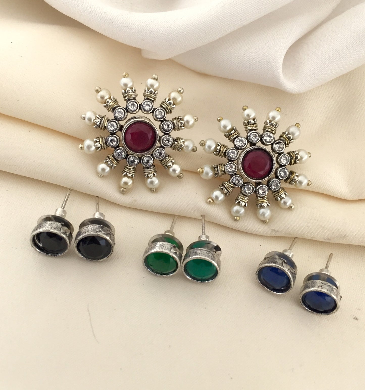 A set of earrings, including a pair of large, ornate earrings with a central red stone surrounded by smaller white and silver stones, and three pairs of smaller stud earrings with black, green, and blue stones.