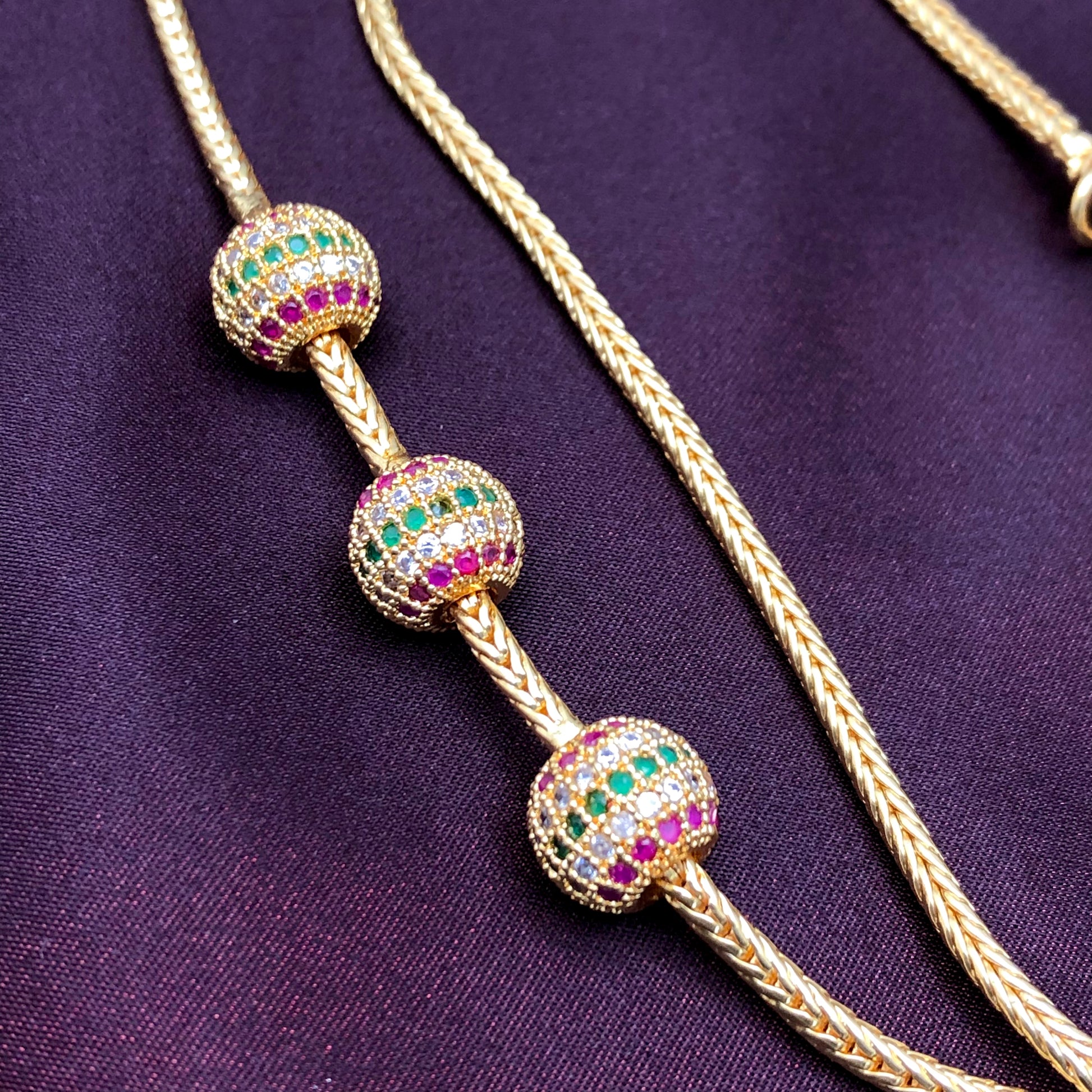 A gold necklace with three spherical beads adorned with green, red, and white gemstones.