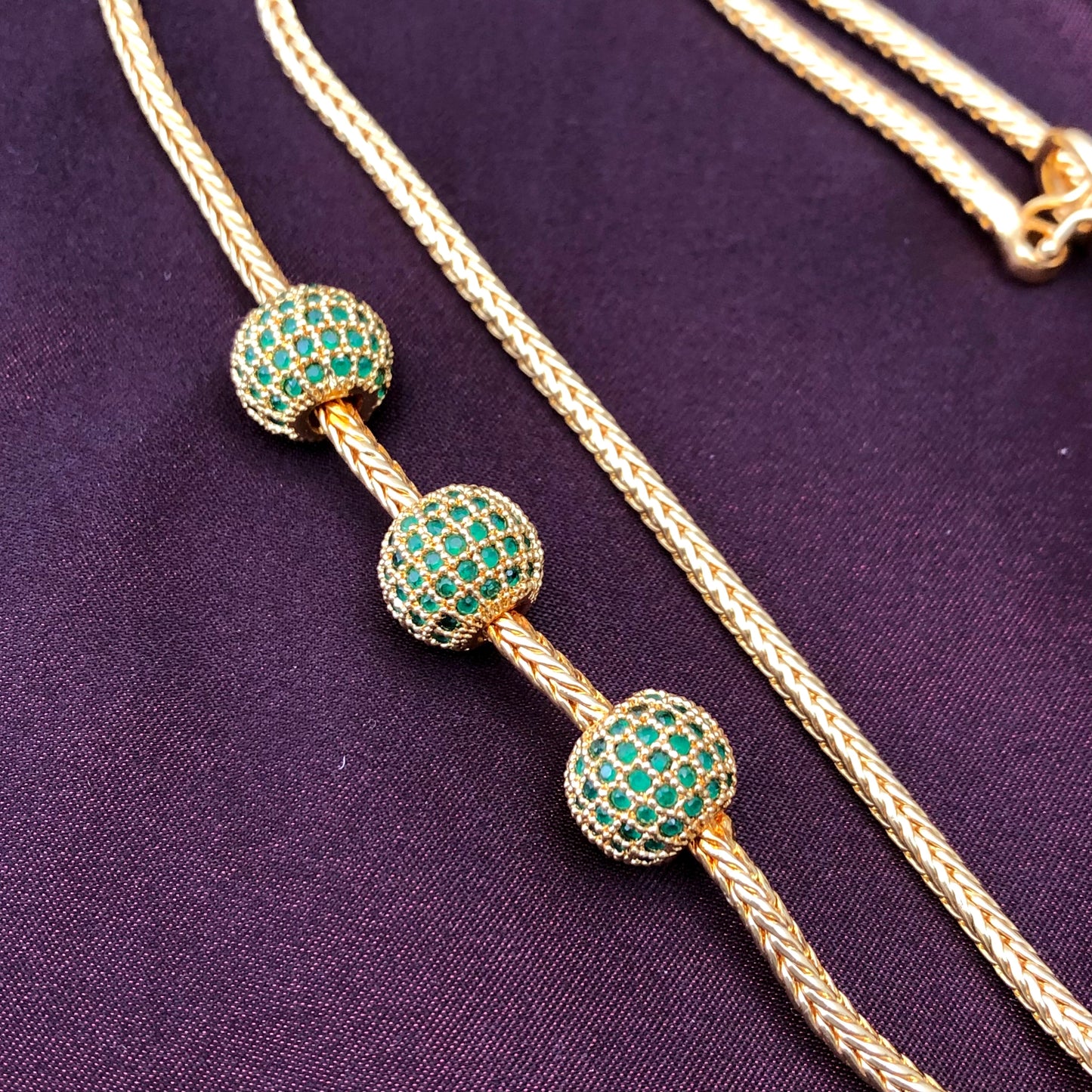 A gold necklace with three green gemstone-studded spherical beads on a dark purple fabric background.