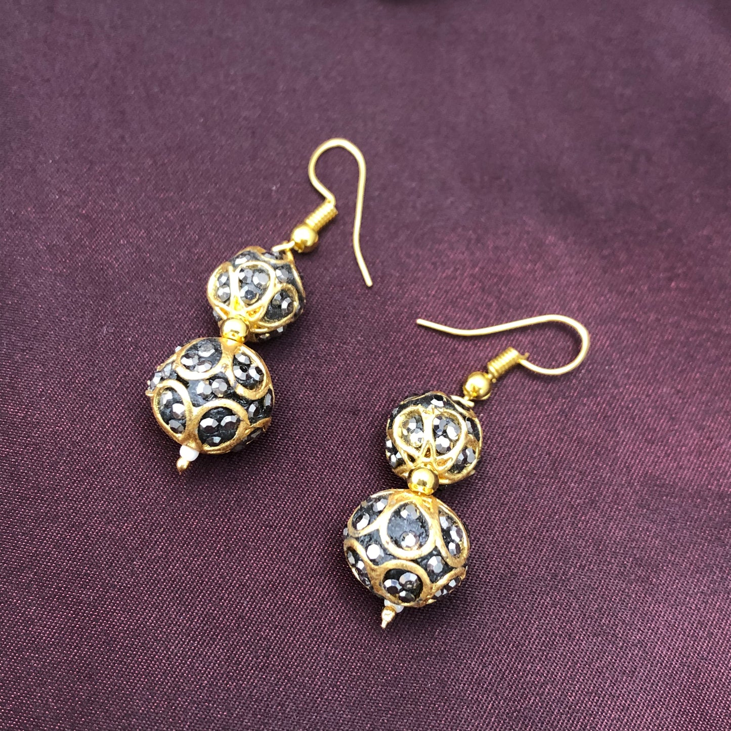 A pair of gold and black beaded drop earrings with intricate designs, placed on a dark purple fabric background.