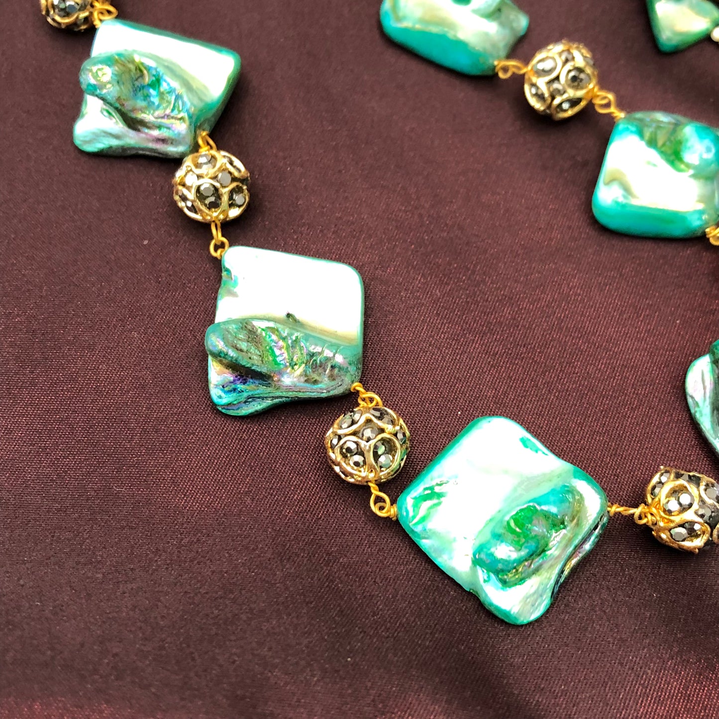 A close-up image of a necklace featuring irregularly shaped, iridescent green beads and ornate gold spherical beads, all linked together with gold wire, displayed on a dark maroon fabric background.