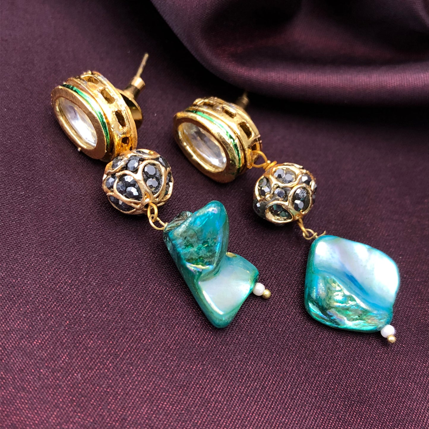 Gold earrings with intricate designs, featuring black gemstone beads and irregularly shaped blue-green stones, displayed on a dark purple fabric background.