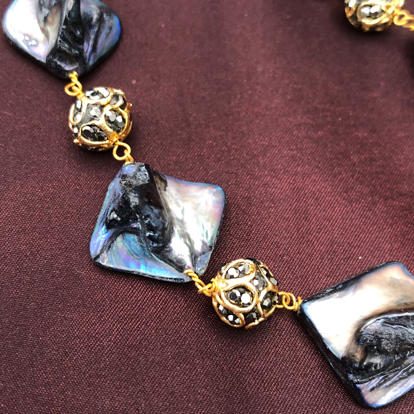 A close-up of a necklace featuring square-shaped, iridescent black and silver beads interspersed with ornate, gold filigree beads, displayed on a dark brown fabric background.
