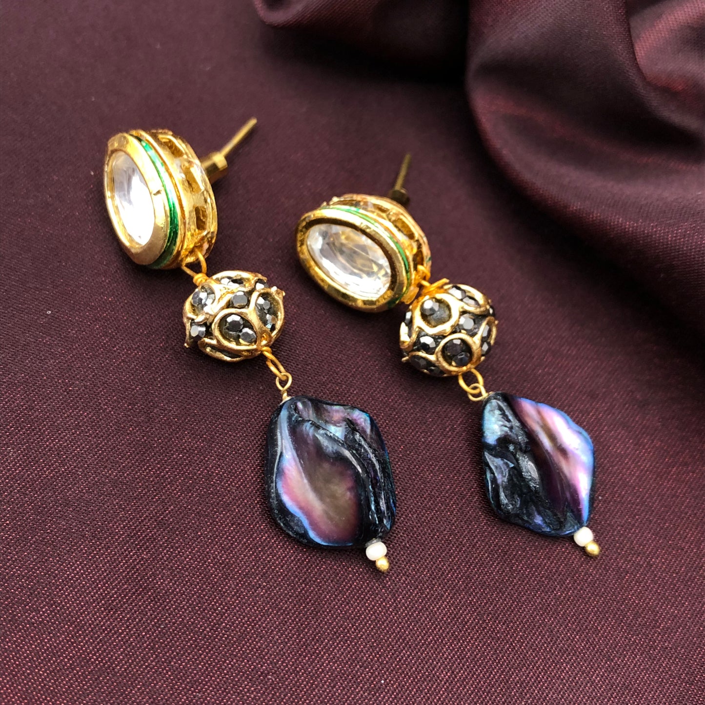 A pair of gold drop earrings with clear gemstones at the top, followed by intricate gold and black bead designs, and finished with iridescent black and purple shell-like pendants. The earrings are displayed on a dark maroon fabric background.