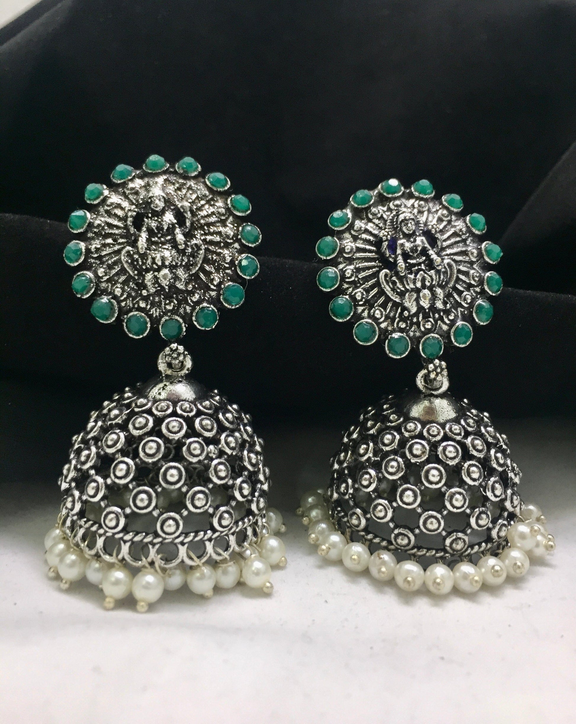A pair of ornate silver jhumka earrings with intricate designs, green stone accents, and pearl embellishments at the bottom.