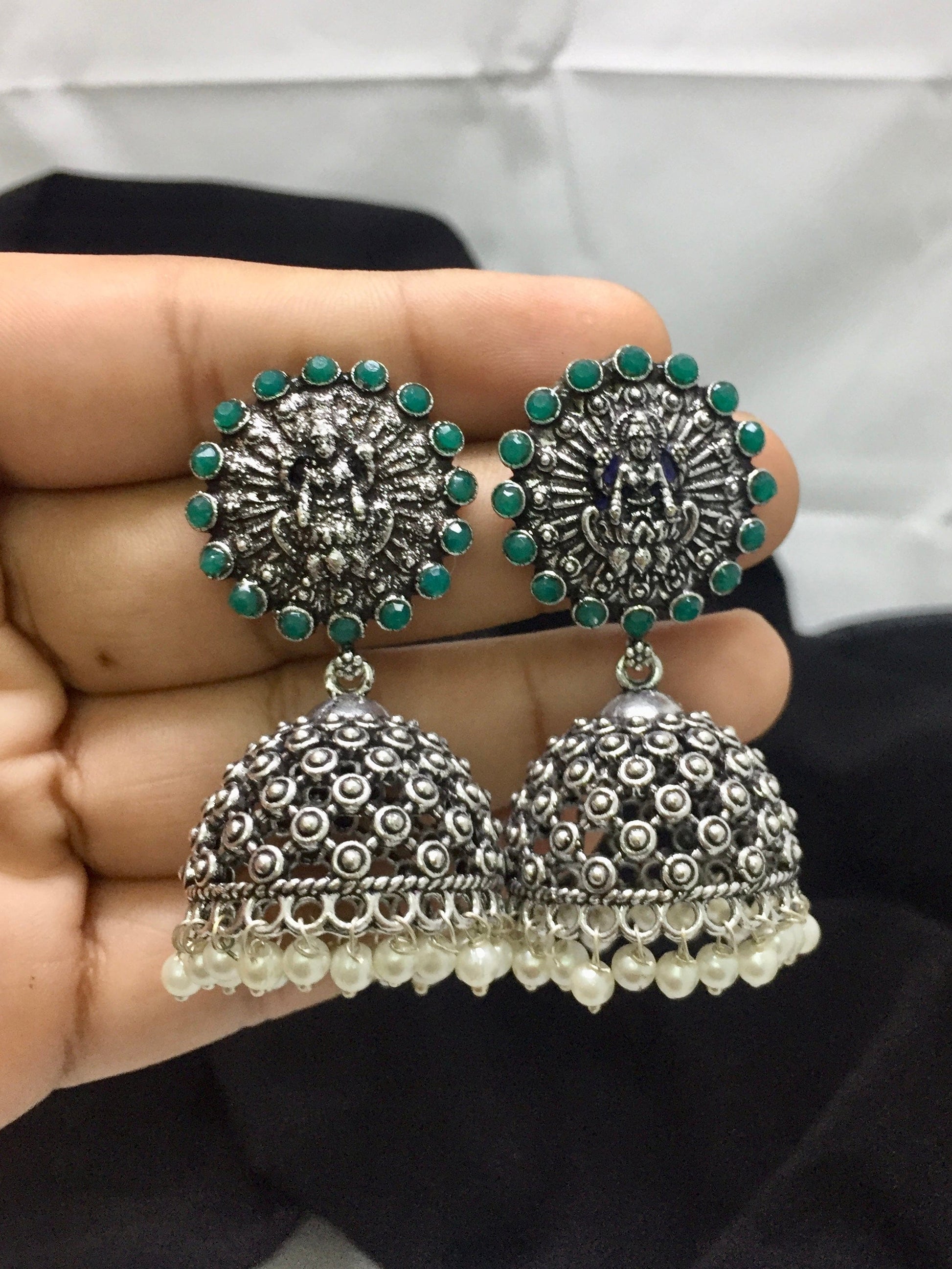 A pair of ornate silver jhumka earrings with green stone accents and pearl detailing.