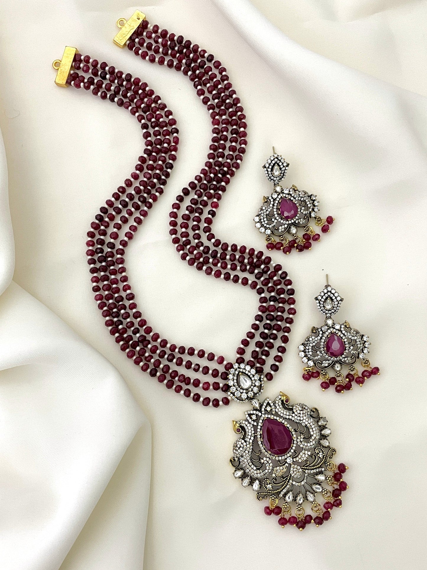 A set of jewelry featuring a multi-strand maroon beaded necklace with an ornate pendant adorned with a large central purple stone and intricate silver detailing, accompanied by matching earrings.