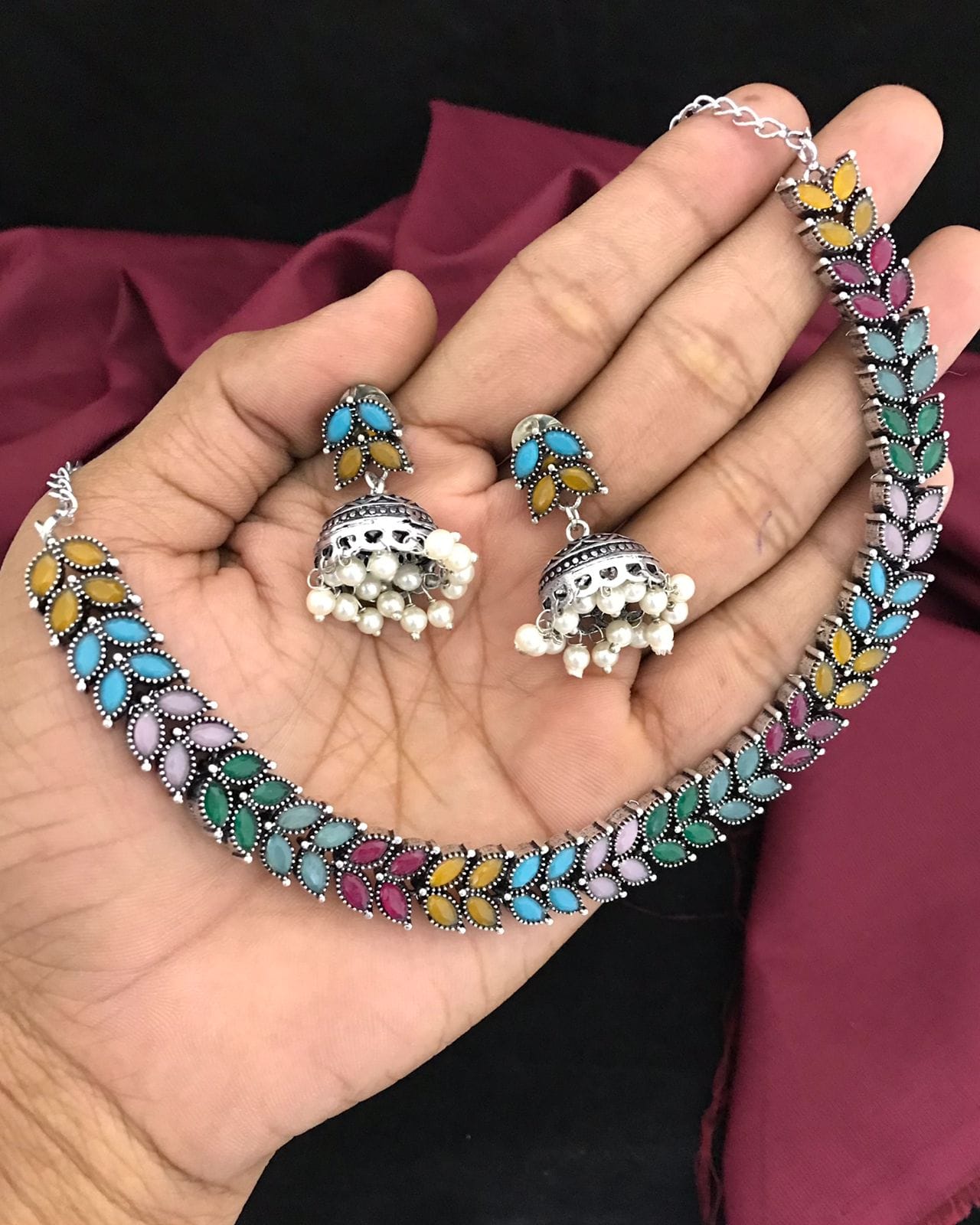 A colorful necklace and matching earrings with intricate designs and pearl accents held in a hand.