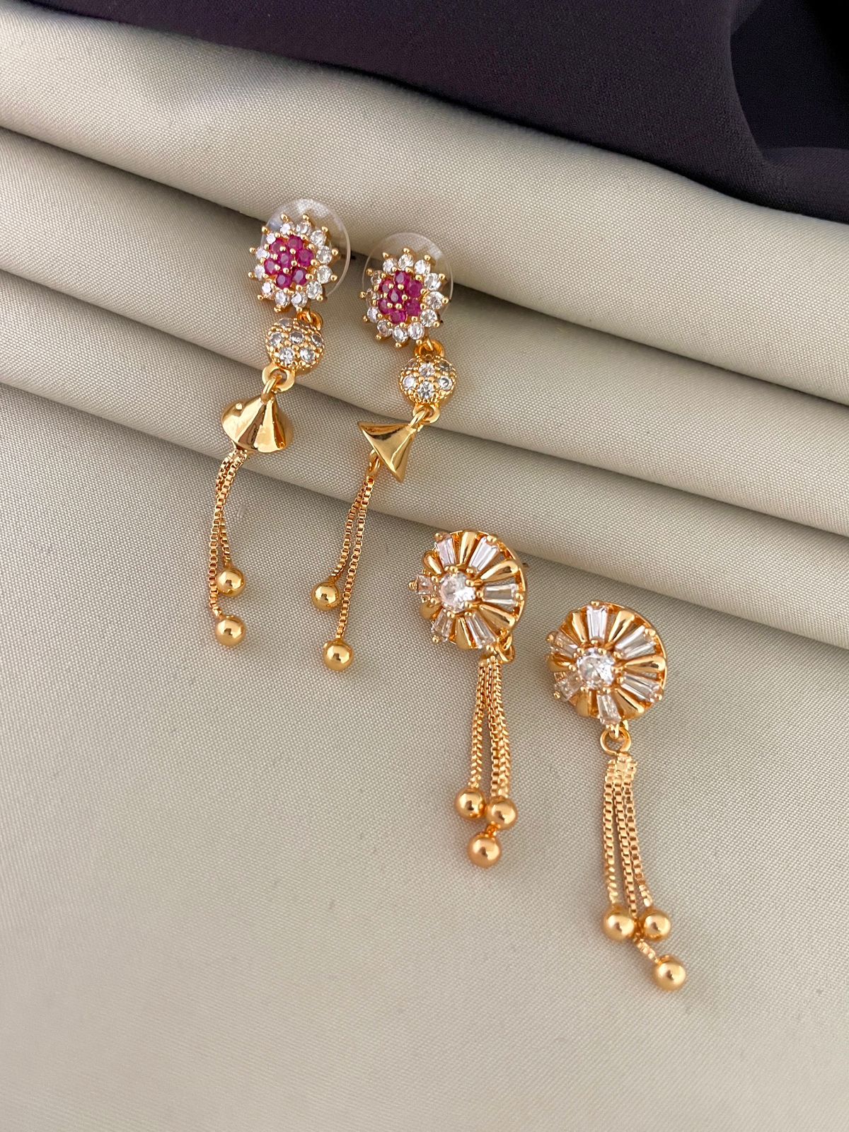 Two pairs of gold earrings with intricate designs and dangling elements. One pair features pink and white stones, while the other pair has white stones arranged in a circular pattern.