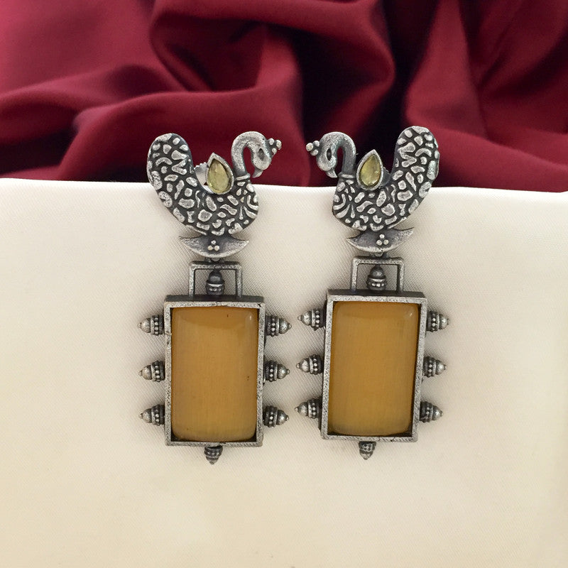 A pair of silver earrings with intricate peacock designs at the top and rectangular amber-colored stones set in ornate frames below, displayed against a white and red fabric background.