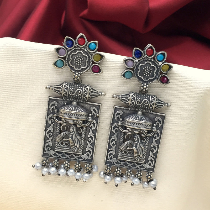 Alt text: A pair of ornate silver earrings with a rectangular base featuring an intricate design and a depiction of a traditional figure. The top part of the earrings is adorned with a flower-like arrangement of colorful gemstones. Small pearl-like beads dangle from the bottom of the earrings.