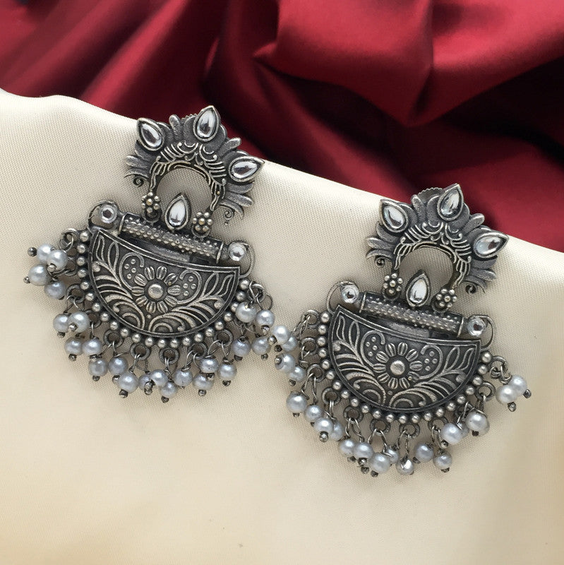 A pair of intricately designed silver earrings with floral patterns and dangling pearl-like beads, displayed on a beige fabric background with a red cloth in the backdrop.