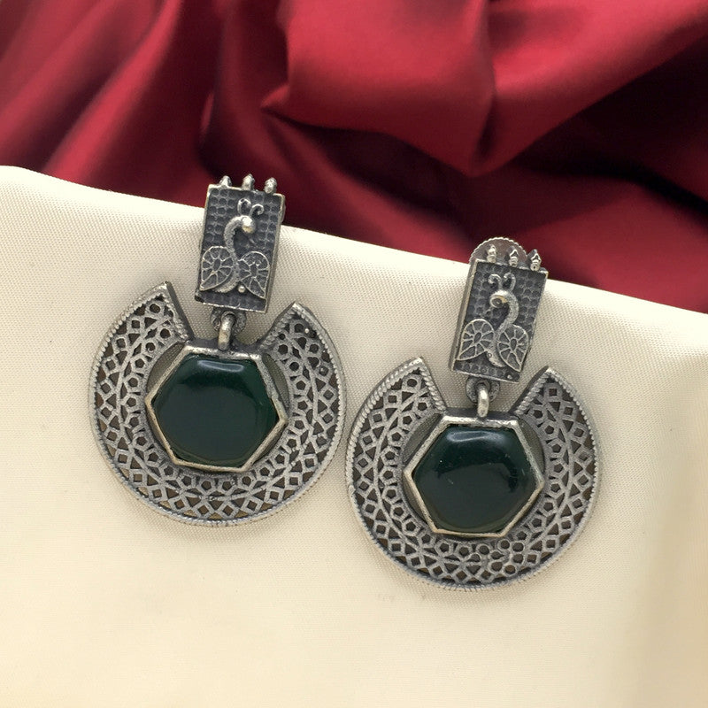 A pair of silver earrings with intricate designs and dark green gemstone centers, displayed against a cream-colored background with a red fabric backdrop.