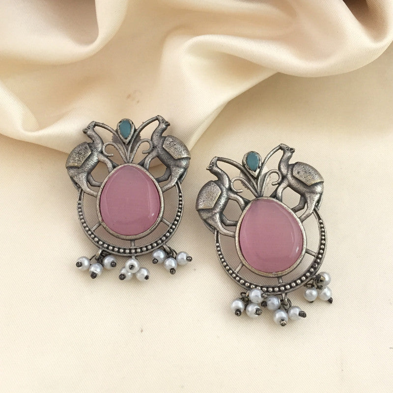 A pair of ornate silver earrings with a large pink stone in the center, surrounded by intricate metalwork and small white pearl-like beads hanging at the bottom. The earrings are placed on a cream-colored fabric background.