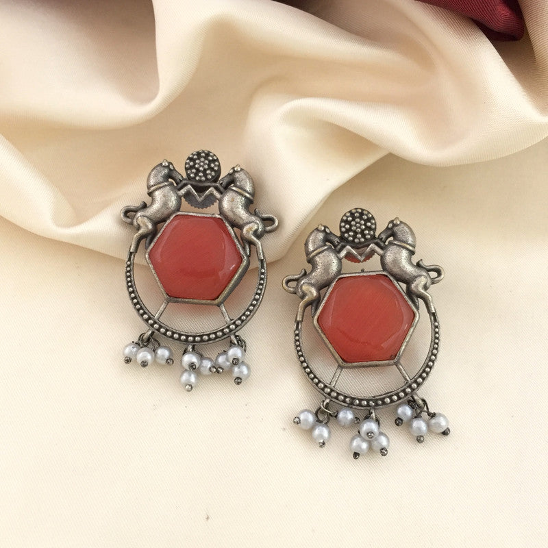 A pair of ornate earrings featuring a hexagonal red gemstone in the center, surrounded by an intricate silver design with two horse figures at the top. The earrings are adorned with small dangling white pearls at the bottom.