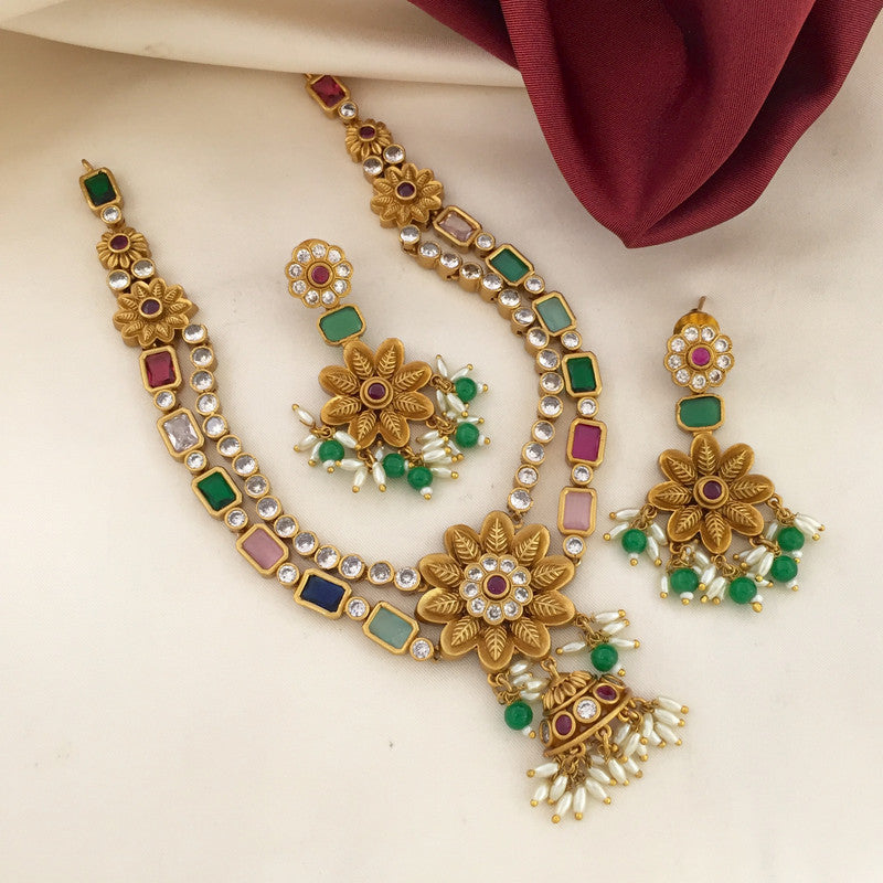 A set of traditional gold-toned jewelry featuring a necklace, a pair of earrings, and a ring. The necklace has floral designs with embedded colorful stones and pearl accents. The earrings and ring also feature similar floral patterns with colorful stones and pearl drops. The jewelry is displayed on a cream-colored fabric with a maroon cloth in the background.