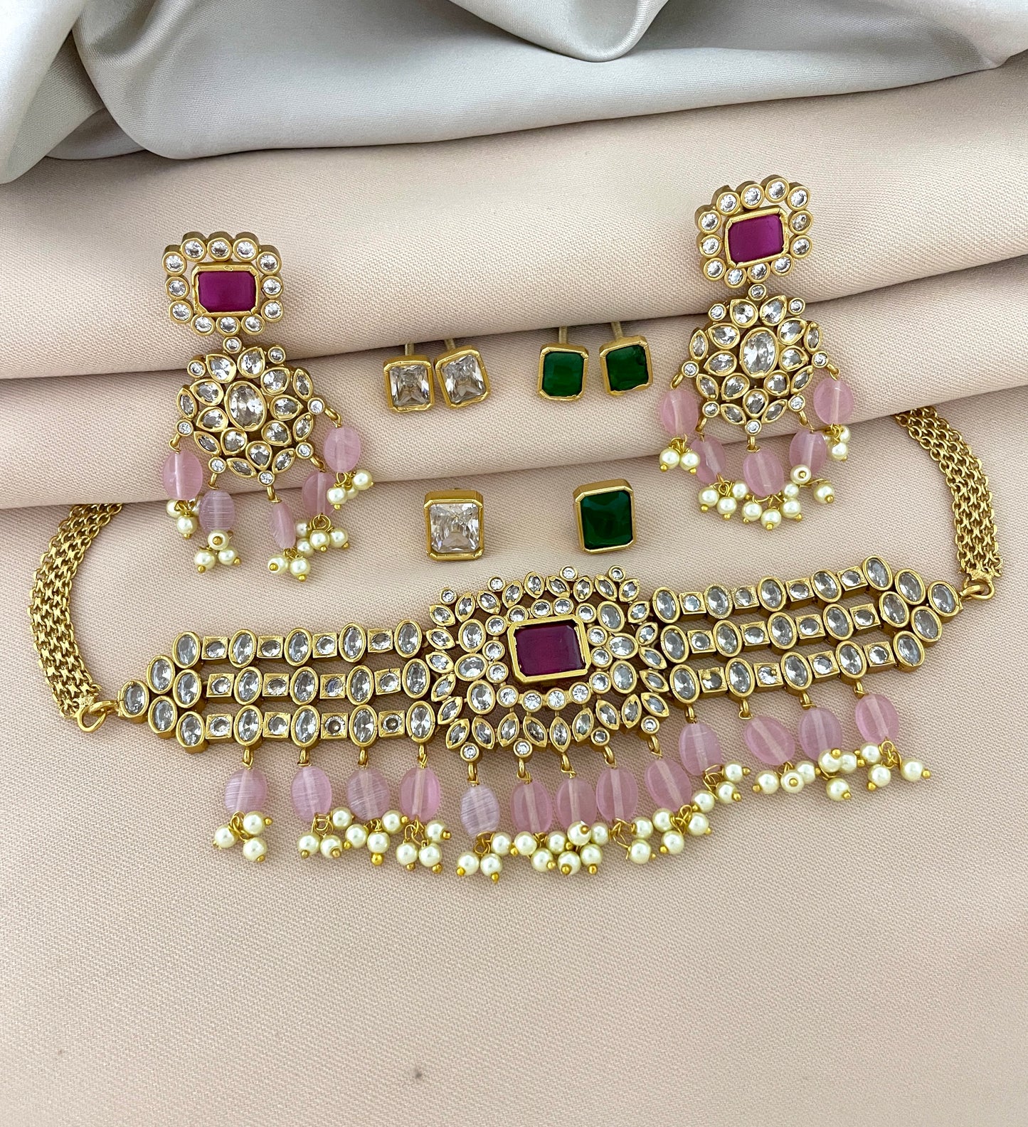 A gold jewelry set featuring a choker necklace with pink and white beads, matching earrings with pink and white beads, and additional stud earrings with green and clear stones.