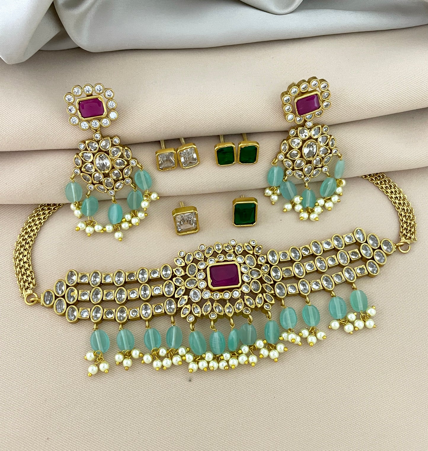 Gold jewelry set with a choker necklace, matching earrings, and stud earrings. The choker features a central red gemstone, surrounded by clear stones and green beads. The matching earrings have a similar design with red gemstones, clear stones, and green beads. The stud earrings include pairs with green and clear stones.